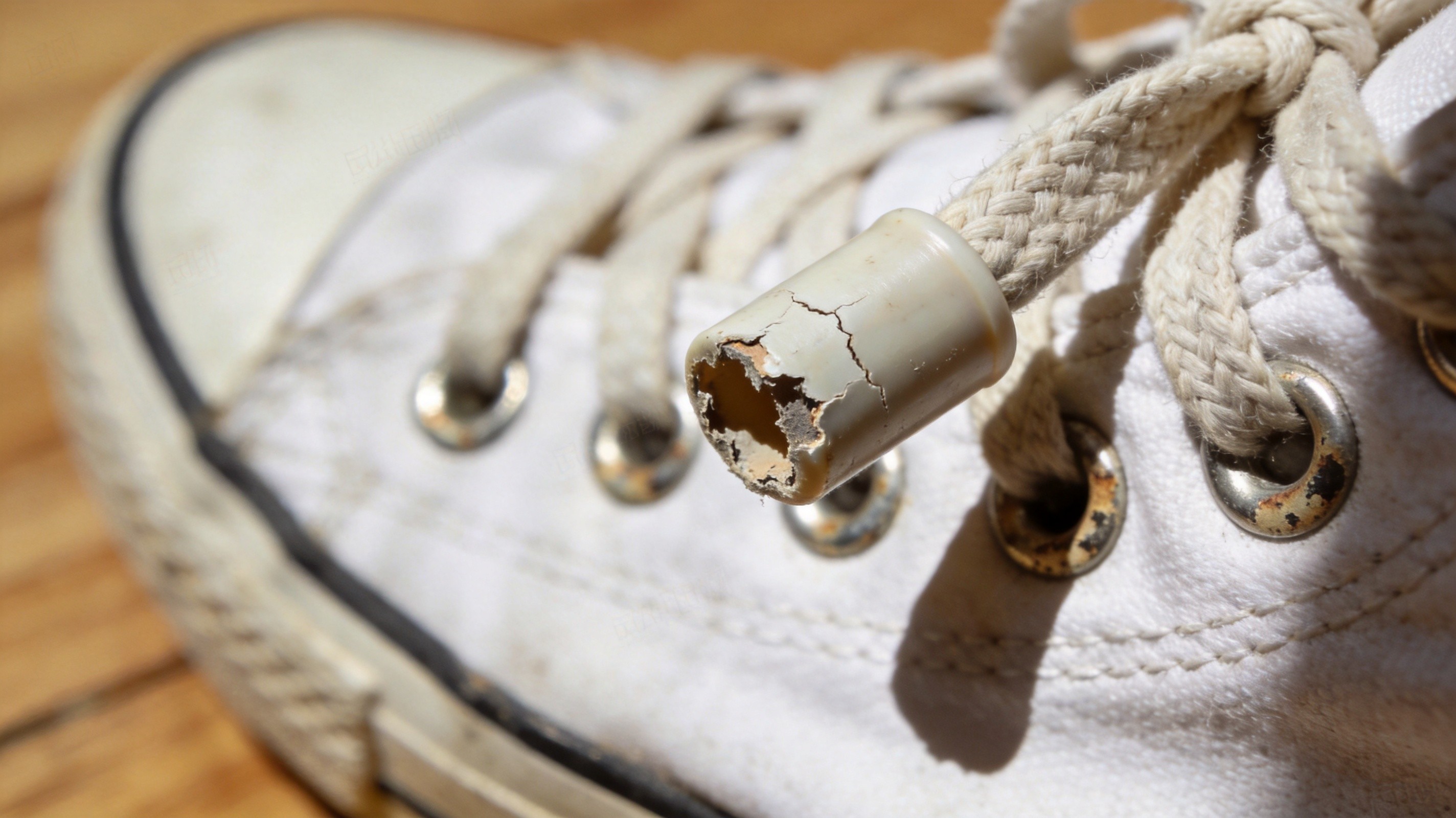 An extreme close-up macro photograph of a cracked and peeling clear plastic aglet on the end of a worn canvas sneaker lace, with a metal eyelet and a small section of white canvas upper visible behind it
